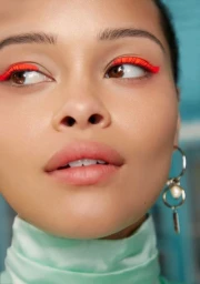 Close-up portrait of a woman wearing bright orange eyeliner and hoop earrings with pearl accents against a blue background.