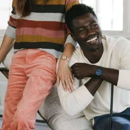 Black man smiling while wearing a white long-sleeve shirt next to a person in a striped sweater and pink trousers.
