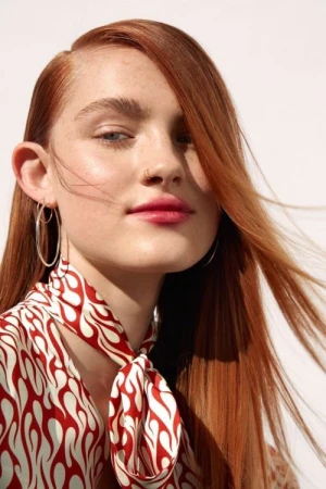Close-up portrait of a woman with red hair and freckles wearing silver hoop earrings and a red and white patterned neck scarf