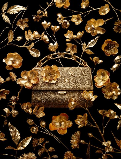 Gold handbag with gold floral embellishments and handle, set against a stark black backdrop, creating a luxurious visual contrast.
