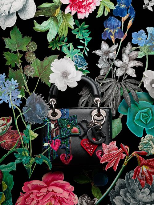 Elegant black handbag with colorful floral embellishments and silver hardware, displayed amidst an arrangement of illustrated flowers on black.