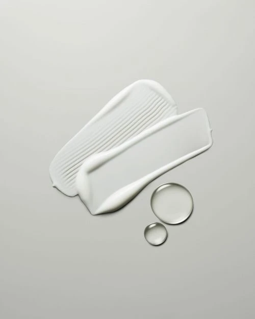 Close-up shot of two textured cream smears and two translucent liquid drops on a gray background.
