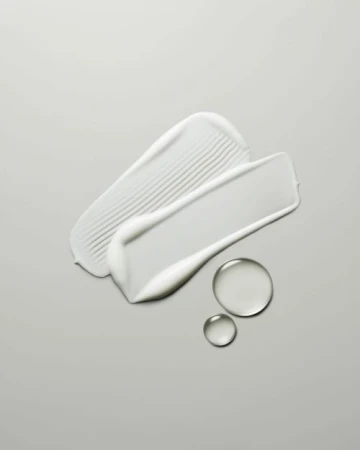 Close-up shot of two textured cream smears and two translucent liquid drops on a gray background.