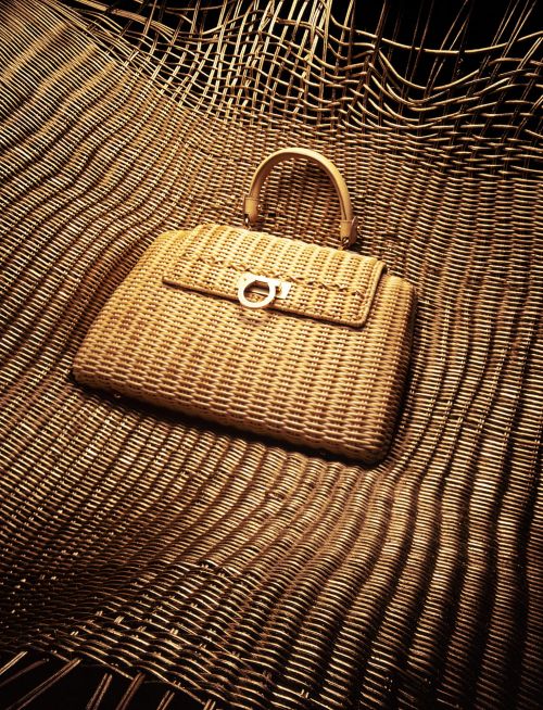 Wicker handbag with light brown handle and clasp placed on a textured, woven surface, shot with warm, even lighting. 