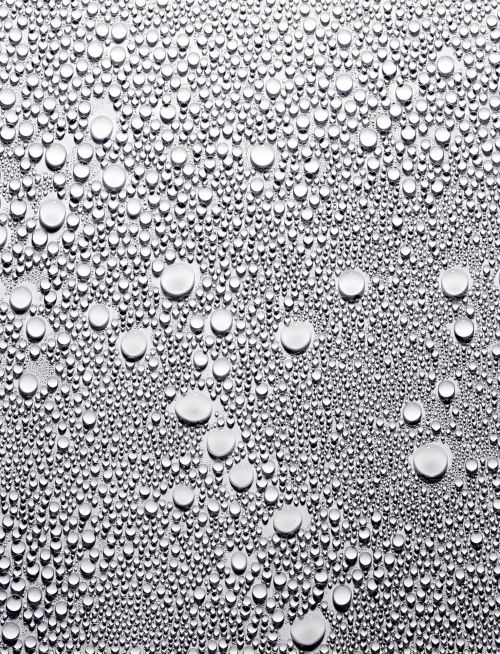 A macro photograph captures countless water droplets of varying sizes scattered across a smooth, silvery metallic surface, creating a textured effect.