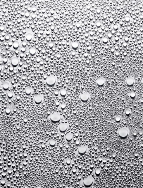 A macro photograph captures countless water droplets of varying sizes scattered across a smooth, silvery metallic surface, creating a textured effect.