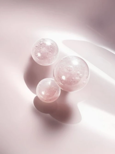 Three translucent pink spheres rest on a smooth, light pink surface, casting soft shadows, suitable for beauty or skincare promotion.