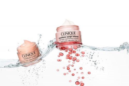 Two jars of Clinique Moisture Surge Intense skin cream stand partially submerged in clear water with coral-colored droplets on white.