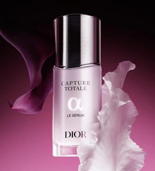 Dior Capture Totale serum bottle with silver cap, set against a backdrop of pink and black, accented by calla lily petals.