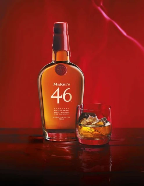 Maker's 46 Kentucky bourbon whiskey bottle next to a glass of whiskey with ice against a red background, studio shot.