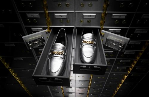 Two silver loafer shoes with gold chains displayed in open drawers of a metal bank vault box unit with drawers, dark neutral lighting.