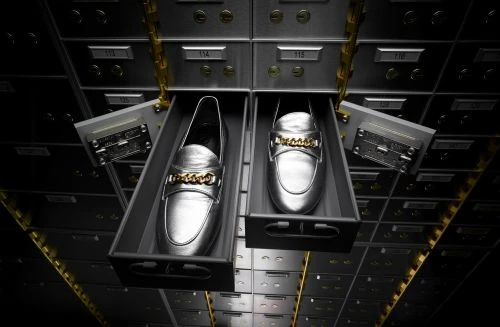 Two silver loafer shoes with gold chains displayed in open drawers of a metal bank vault box unit with drawers, dark neutral lighting.