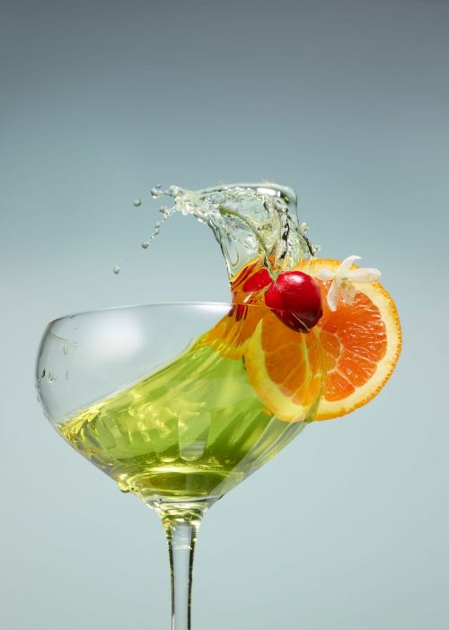 A vibrant cocktail in a stemmed glass, garnished with orange slices, cherries, and water splash, against a light blue background.