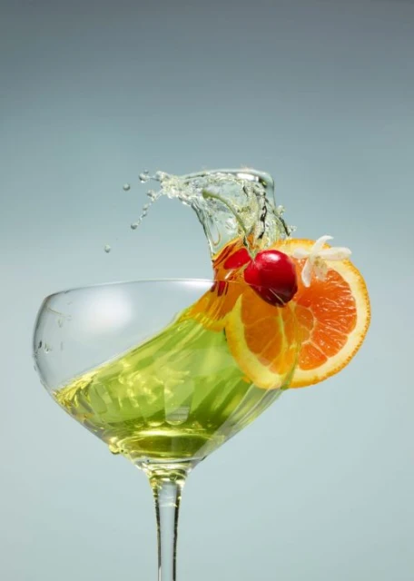 A vibrant cocktail in a stemmed glass, garnished with orange slices, cherries, and water splash, against a light blue background.