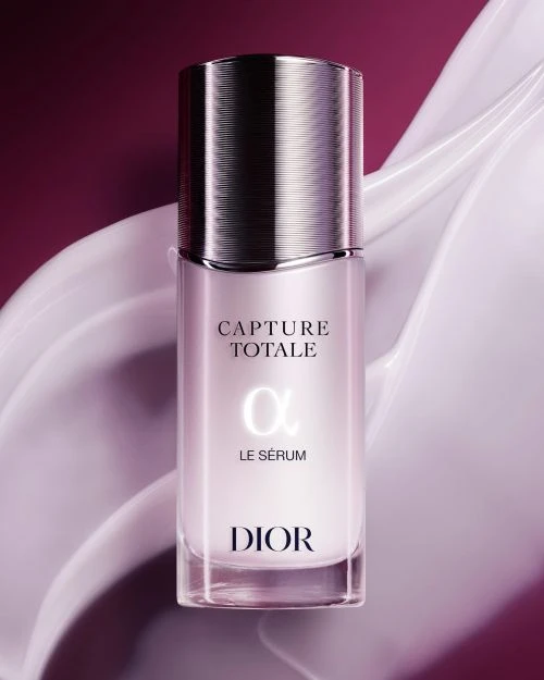 Dior Capture Totale serum in a clear glass bottle with a silver lid on a pink background with white cream swirls, beauty shot.