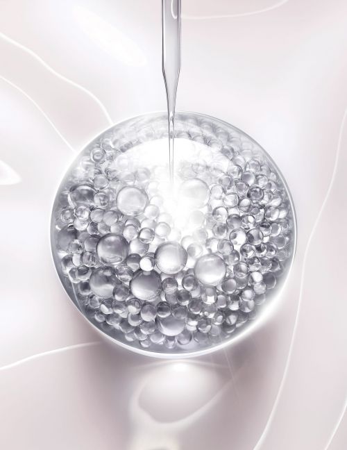 Close-up of clear serum droplet falling onto a translucent sphere filled with smaller silver beads against a pale pink background.