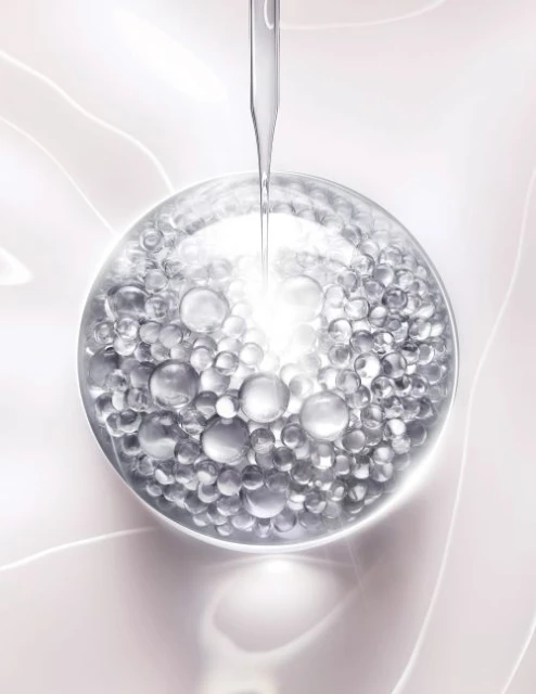 Close-up of clear serum droplet falling onto a translucent sphere filled with smaller silver beads against a pale pink background.