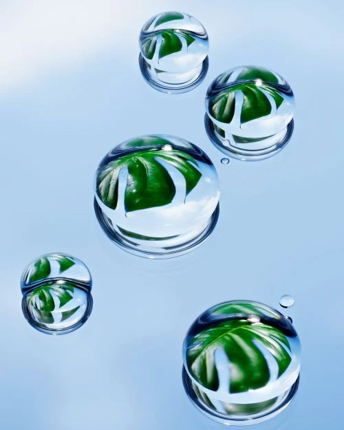 Macro photography of water droplets with green leaves reflected inside, set against a soft blue gradient background.