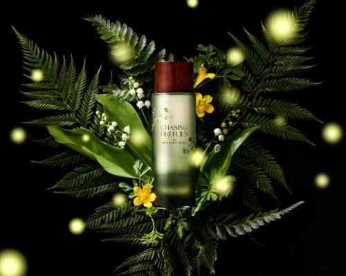 Still life of perfume bottle with brown wood cap surrounded by green ferns, small white and yellow flowers, and glowing firefly lights.