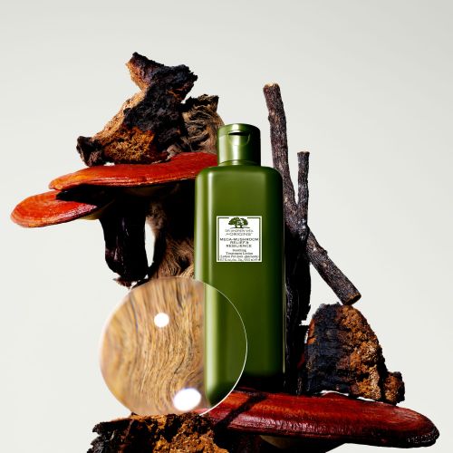Green Origins skincare bottle surrounded by natural elements like mushrooms, wood, and a clear acrylic disc on an off-white background.