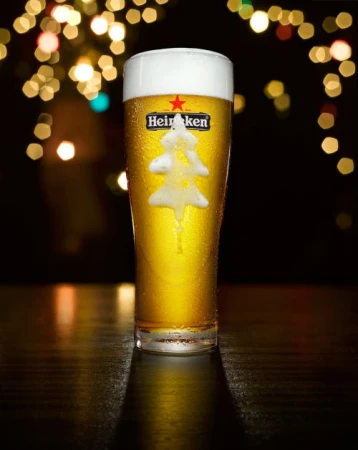 Frothy glass of Heineken beer with a tree-shaped foam design, set against a dark background with bokeh lights. Festive drink.