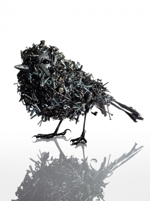 Sculptural bird made of screws, bolts, and metal pieces standing in photo studio. Black bird with white background and reflection.