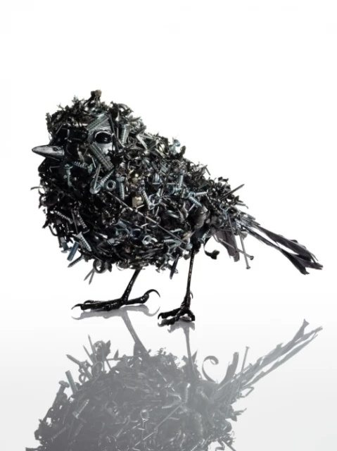 Sculptural bird made of screws, bolts, and metal pieces standing in photo studio. Black bird with white background and reflection.