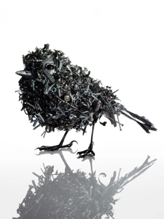 Sculptural bird made of screws, bolts, and metal pieces standing in photo studio. Black bird with white background and reflection.