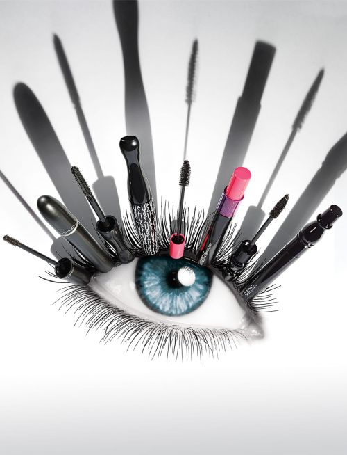 Creative makeup concept featuring a human eye surrounded by mascara wands and a lipstick, shot in a photo studio with soft lighting.