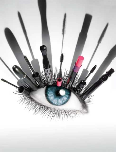 Creative makeup concept featuring a human eye surrounded by mascara wands and a lipstick, shot in a photo studio with soft lighting.