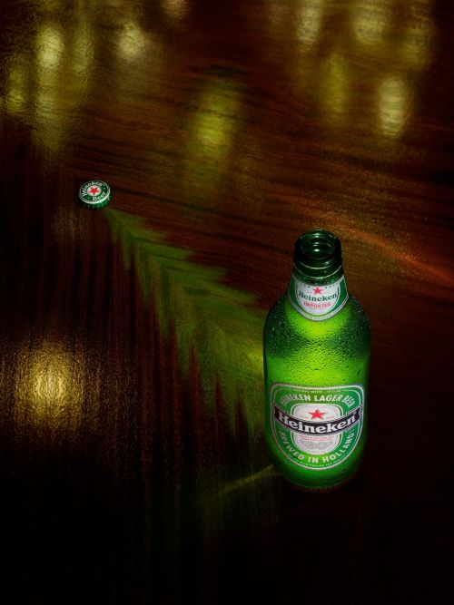 A close-up studio shot showcasing a green Heineken beer bottle with condensation, positioned next to its cap on a dark wooden surface.