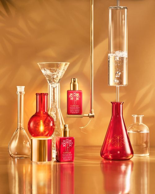 Clinique skincare product bottles with gold accents are arranged with glass beakers on a glossy gold surface against a gold background.
