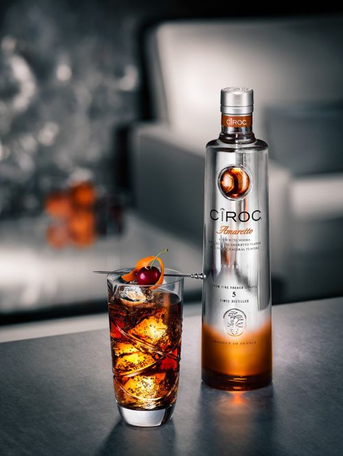 Ciroc Amaretto vodka bottle and cocktail drink with ice, cherry, and orange peel garnish on a table in a studio setting.
