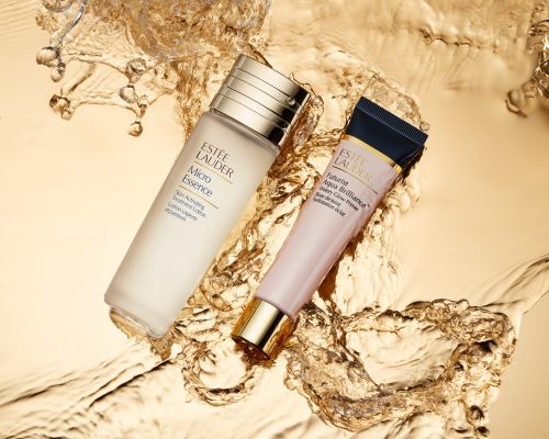 Two Estee Lauder skincare products, Micro Essence and Futurist Aqua Brilliance primer, displayed against a dynamic splash of gold liquid.