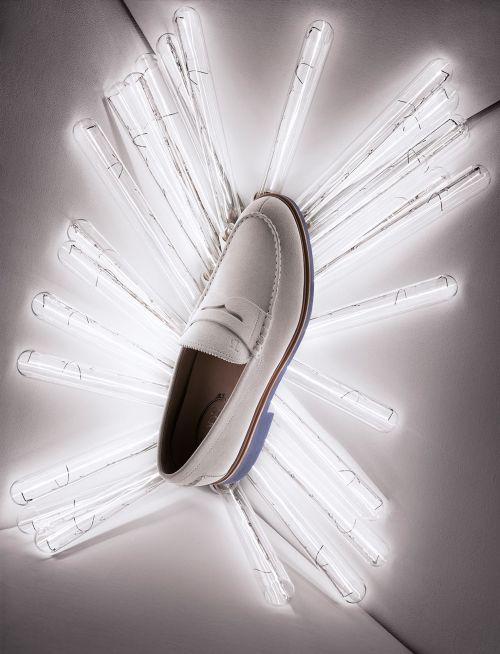 Studio shot of white leather penny loafer surrounded by illuminated glass rods creating a starburst effect on a neutral gray background.