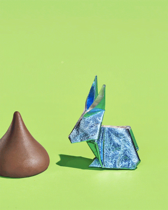 Close-up shot of a brown chocolate kiss candy beside a shiny blue origami rabbit on a light green background with soft studio lighting.