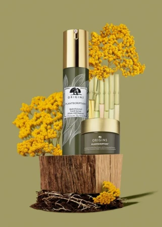 Two Origins Plantscription anti-aging skincare products, a serum and moisturizer, are stacked on a textured wooden block with yellow flowers.
