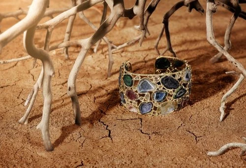 Gold cuff bracelet with colorful gemstones on a cracked earth surface surrounded by dried branches, jewelry product shot.