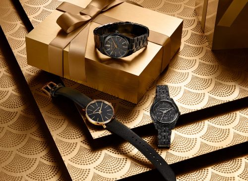Still life of three watches, one on a gold gift box with a ribbon, on a patterned gold surface with Art Deco-style fan shapes.