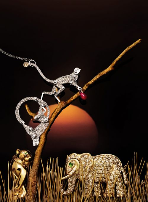 Jewelry still life features diamond encrusted gold elephant, monkey, and lion animal pendants against a dark background with a sunset.