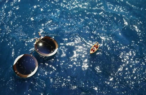 Aerial view of a small boat with people on a sparkling blue body of water, next to two floating platforms with solar panels.