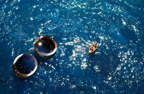 Aerial view of two circular structures floating on a blue sea with a small boat containing two people, reflecting sunlight.