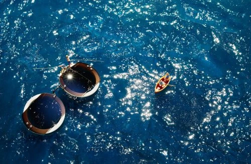 Aerial view of two circular structures floating on a blue sea with a small boat containing two people, reflecting sunlight.