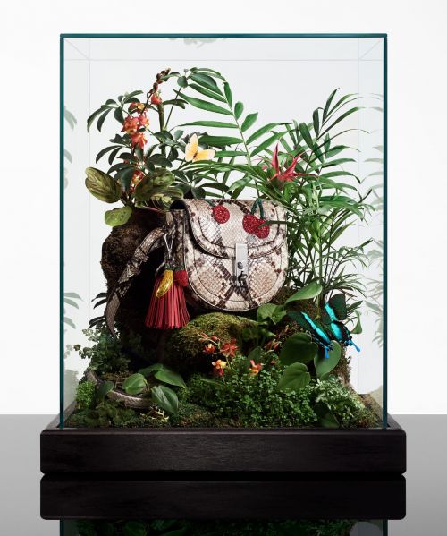Fashion handbag with cherry accents in a glass display case surrounded by tropical foliage, creating a miniature jungle scene.