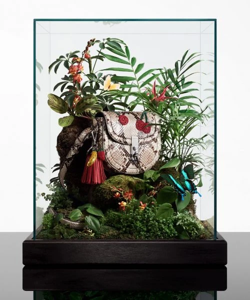 Fashion handbag with cherry accents in a glass display case surrounded by tropical foliage, creating a miniature jungle scene.