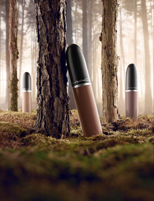 Cosmetic product shot: A row of brown lipsticks by MAC stands in a forest setting. The backdrop features blurred trees and mossy ground.