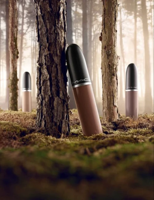 Cosmetic product shot: A row of brown lipsticks by MAC stands in a forest setting. The backdrop features blurred trees and mossy ground.