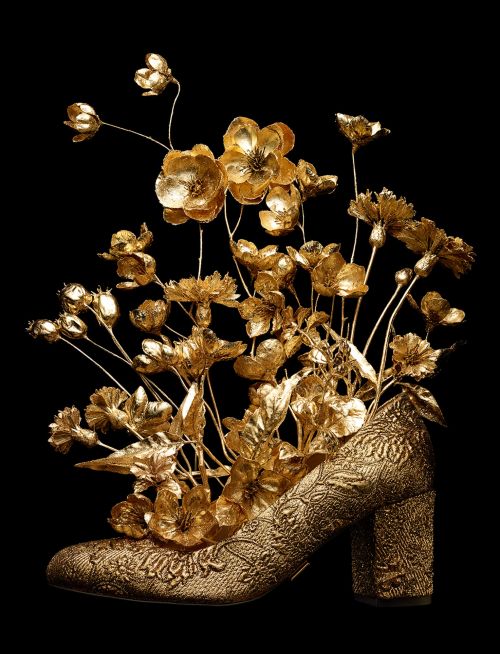 Gold high-heel pump with flowers and stems arranged inside, set against a solid black background, metallic sheen, ornate details.