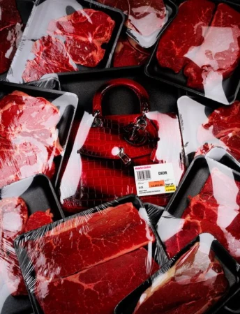 Overhead studio shot of a red leather handbag placed among several packages of raw meat wrapped in plastic and arranged on a black surface.