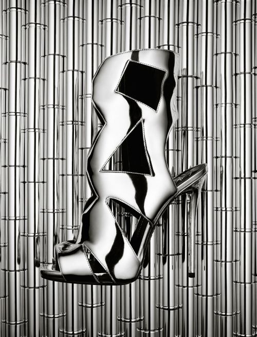 High-heeled silver sandal with geometric cutouts set against a backdrop of shiny silver cylinders, creating a visually striking fashion image.